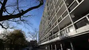 A low-angle view of the Riverview Condominium building in Cambridge, showing its mid-century modern architecture with white concrete framing, balconies, and a row of parked cars underneath. Bare tree branches stretch across a clear blue sky, capturing the stillness of a cold day.